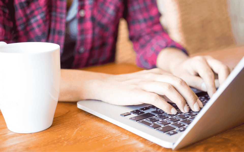 Person's hands typing on a laptop next to a white mug